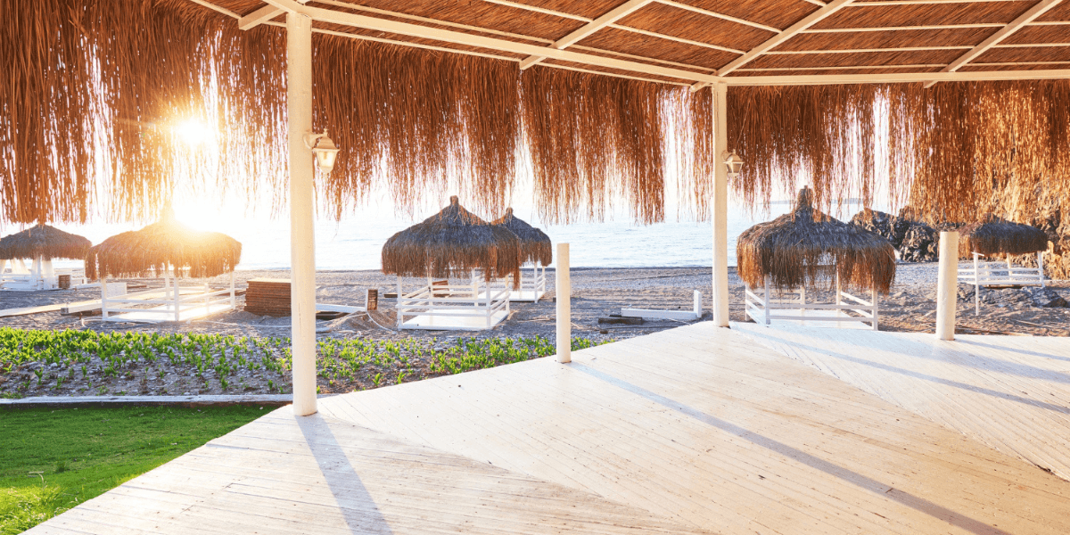 A beach gazebo with thatched umbrellas providing shade, surrounded by sand and ocean waves in the background.